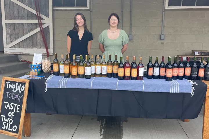 Two people stand behind a table with various wine bottles and a sign for a wine tasting.
